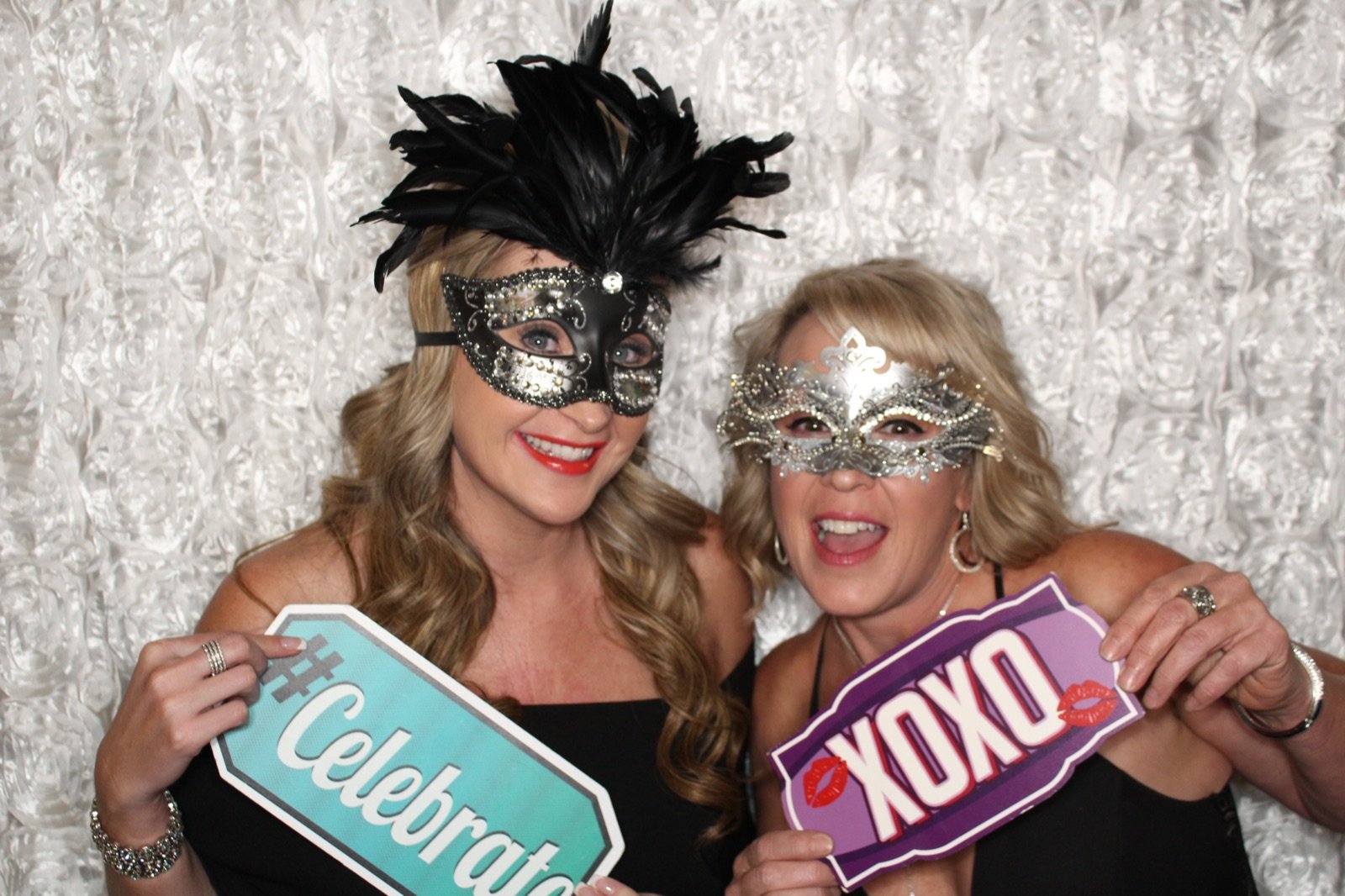 Guests in a black feather mask and silver mask holding Celebrate and XOXO photo booth signs